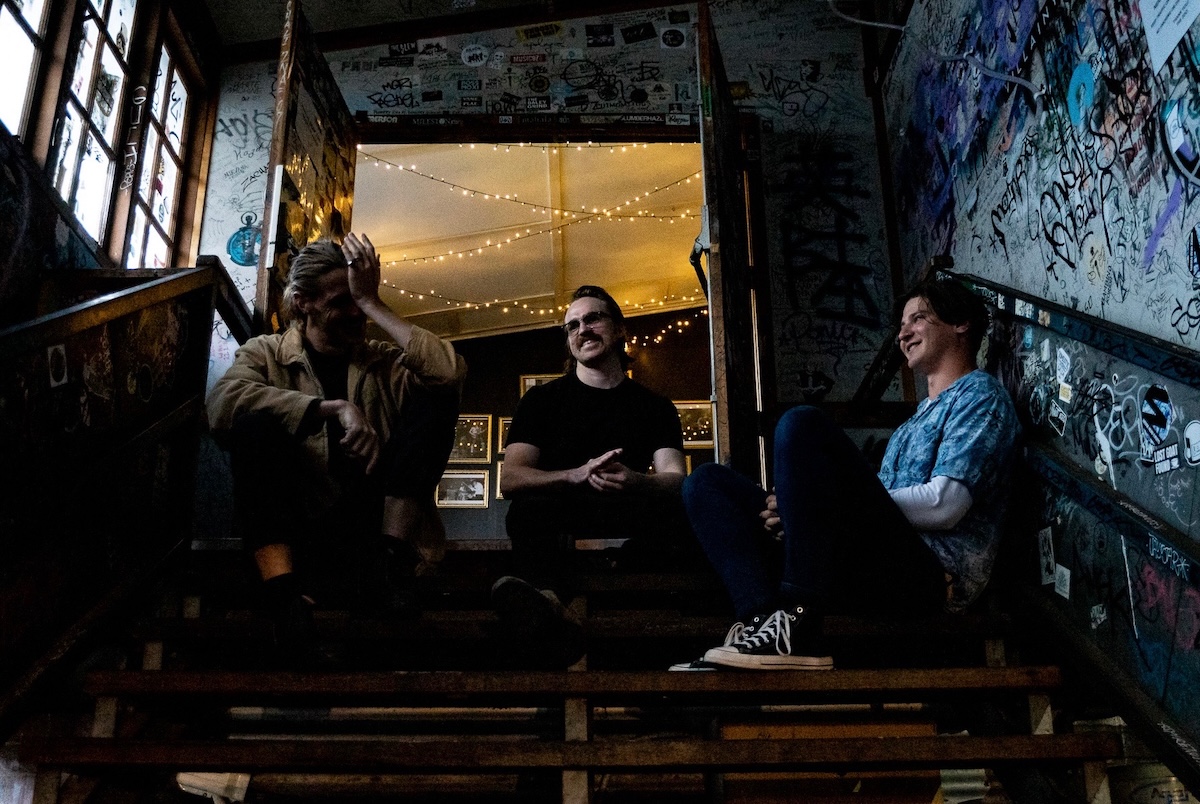 Brisbane indie rock band Bugs sitting on a graffiti-covered staircase inside a music venue, laughing together during a promo shot.
